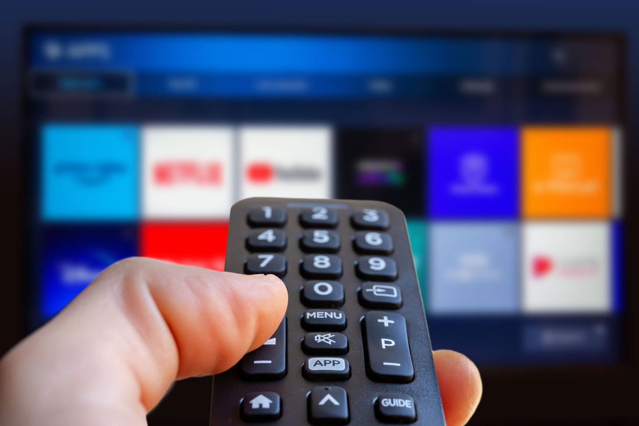 A man is holding a remote control of a smart TV in his hand. In the background you can see the television screen with streaming entertainment apps for video on demand